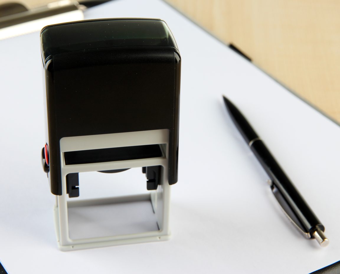 Self-inking stamp and a pen resting on a blank sheet on a light wooden desk
